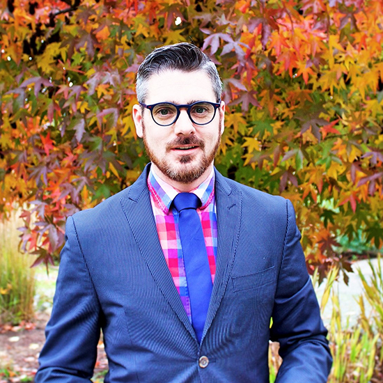 Headshot of a man with black glasses, a moustache beard, and wearing a blue blazer and tie over a checkered shirt